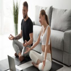 Smiling millennial couple practice yoga on mats at home watching video tutorial on laptop, excited man and woman sit in lotus position meditating repeating online instructions by coach on computer